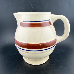 May include: A cream-colored ceramic pitcher with a visible crackle glaze finish. The pitcher features two horizontal bands of dark red paint, each bordered by thin, dark blue stripes encircling the rounded body and the upper neck area.
