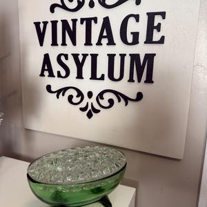 May include: A green glass flower frog with a clear glass top, on a white surface. The sign in the background reads "VINTAGE ASYLUM" in black letters with decorative flourishes.