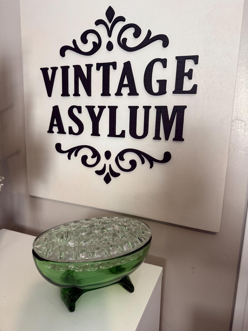 May include: A green glass flower frog with a clear glass top, on a white surface. The sign in the background reads "VINTAGE ASYLUM" in black letters with decorative flourishes.