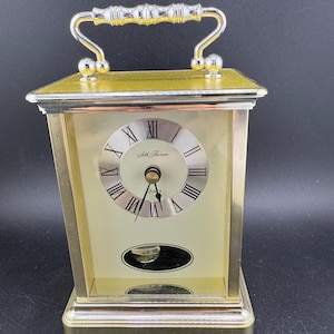 Może przedstawiać: Złoty zegar kwarcowy Seth Thomas z uchwytem. Tarcza zegara ma rzymskie cyfry i nazwę marki. Zegar ma prostokątny kształt z oknem wahadła i ozdobnym uchwytem.