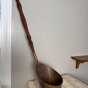 May include: A vintage copper and brass ladle with a long, dark wooden handle. The ladle has a rounded, oval-shaped bowl and a brass band around the base. The handle is attached with a brass fitting. The ladle is leaning against a white wall.