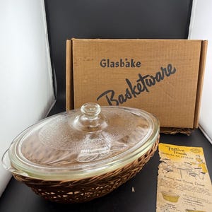 May include: A Glasbake Basketware baking dish with a clear glass lid and a woven basket base. The dish is oval-shaped and comes with its original cardboard box and a product information card.
