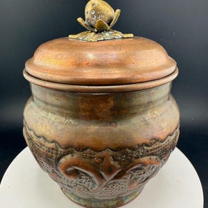 May include: An antique copper jar with a lid, featuring embossed floral patterns and a decorative finial. The finial is an egg-shaped design with a flower base. The jar's body has a textured, aged appearance. The jar is displayed on a white surface.