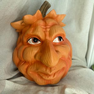 May include: A decorative orange pumpkin sculpture featuring a carved human-like face with a prominent nose, deep-set eyes, and a textured surface. The pumpkin has a brown stem at the top and is shaped with deep ridges, creating a detailed facial expression.