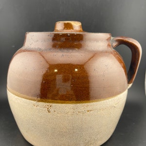 May include: A brown and white glazed ceramic crock with a lid and a handle. The crock has a brown top and a white bottom.