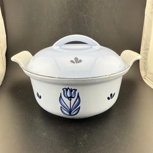 Puede incluir: Una cacerola de metal esmaltado en color azul claro con tapa a juego. El recipiente presenta una ilustración de un tulipán azul oscuro en la parte frontal, pequeños detalles de hojas en azul oscuro y dos asas laterales con textura estriada.