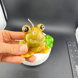 May include: A frog-shaped candle in shades of yellow and green. The candle has large, detailed eyes and a small, green frog candle next to it. The candle is held by a hand.