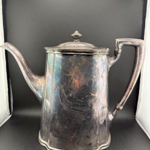 Puede incluir: Una tetera vintage de color plateado con tapa y boquilla curva. La tetera tiene un asa y una base decorativa. La superficie muestra signos de desgaste, lo que indica su antigüedad. La tetera es probablemente de metal y está diseñada para servir bebidas calientes.