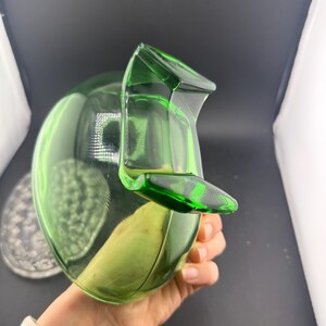 May include: A green glass jug with a rounded body and a spout. The jug is held in a hand, and the background is a dark grey. The jug is approximately 20 cm tall.