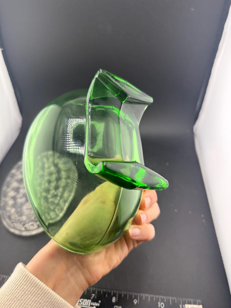 May include: A green glass jug with a rounded body and a spout. The jug is held in a hand, and the background is a dark grey. The jug is approximately 20 cm tall.