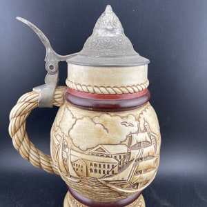 May include: A ceramic beer stein with a pewter lid and a rope handle. The stein features a detailed scene of a sailing ship in a harbor with buildings in the background. The text "Headed Ketch & Barque" is inscribed on the base.