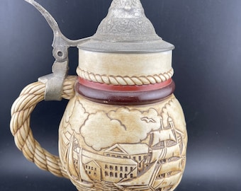 May include: A ceramic beer stein with a pewter lid and a rope handle. The stein features a detailed scene of a sailing ship in a harbor with buildings in the background. The text "Headed Ketch & Barque" is inscribed on the base.