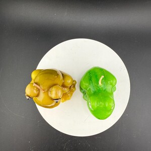 May include: Two decorative candles on a white circular surface. One candle is yellow and shaped like a seated figure with round orbs. The other candle is green and shaped like a pepper. The background is black.