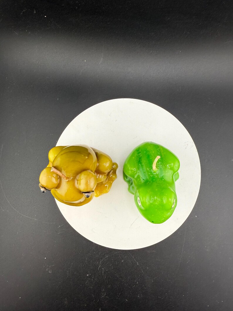 May include: Two decorative candles on a white circular surface. One candle is yellow and shaped like a seated figure with round orbs. The other candle is green and shaped like a pepper. The background is black.