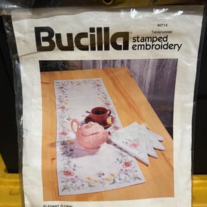 May include: A Bucilla stamped embroidery kit for a tablerunner and set of 8 napkins. The packaging features a photo of a white tablerunner with a floral border, a pink teapot, and a cup on a wooden table. Text includes 40714 and ELEGANT FLORAL.