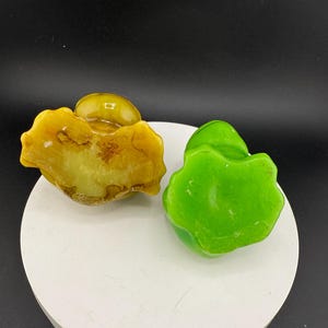 May include: Two decorative stone sculptures. One is a light yellow colour with brown marbling, and the other is a vibrant green. Both have a rounded, organic shape, and are displayed on a white surface against a black background.