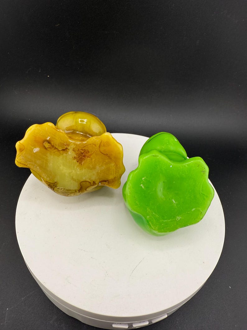 May include: Two decorative stone sculptures. One is a light yellow colour with brown marbling, and the other is a vibrant green. Both have a rounded, organic shape, and are displayed on a white surface against a black background.
