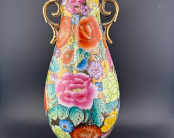 May include: A colorful, hand-painted ceramic vase with a floral design. The vase features a variety of flowers in shades of pink, orange, yellow, and blue. It has two gold-colored handles and a gold rim. The vase is approximately 10 inches tall.