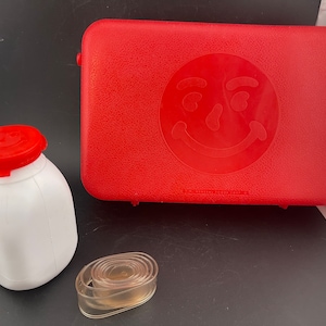 May include: A red plastic lunchbox with a smiley face design. The lunchbox is closed and has a white plastic container and a clear plastic band next to it.