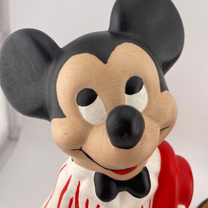 Puede incluir: Una figurita de cer&aacute;mica de Mickey Mouse con una camisa a rayas blancas y rojas, una pajarita negra y un sombrero rojo. Est&aacute; sosteniendo un objeto rojo en su mano derecha.