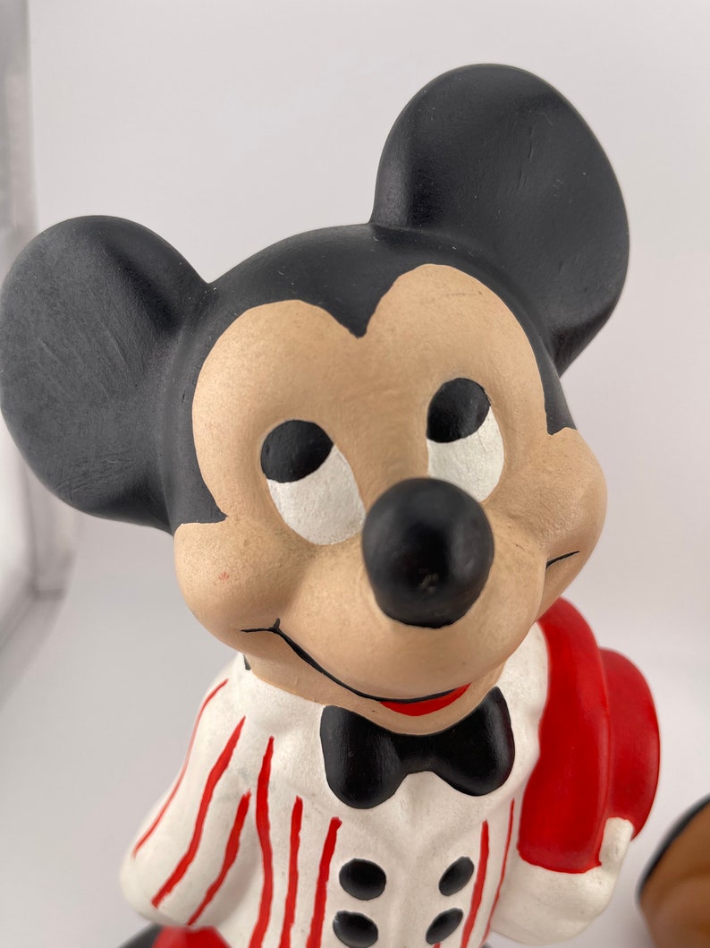 Puede incluir: Una figurita de cer&aacute;mica de Mickey Mouse con una camisa a rayas blancas y rojas, una pajarita negra y un sombrero rojo. Est&aacute; sosteniendo un objeto rojo en su mano derecha.