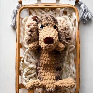 May include: A handmade crochet dog toy in a wooden basket, surrounded by white yarn and string lights. The dog is brown and tan with a dark red nose. The words "YOU ARE LOVED" are displayed above the basket.