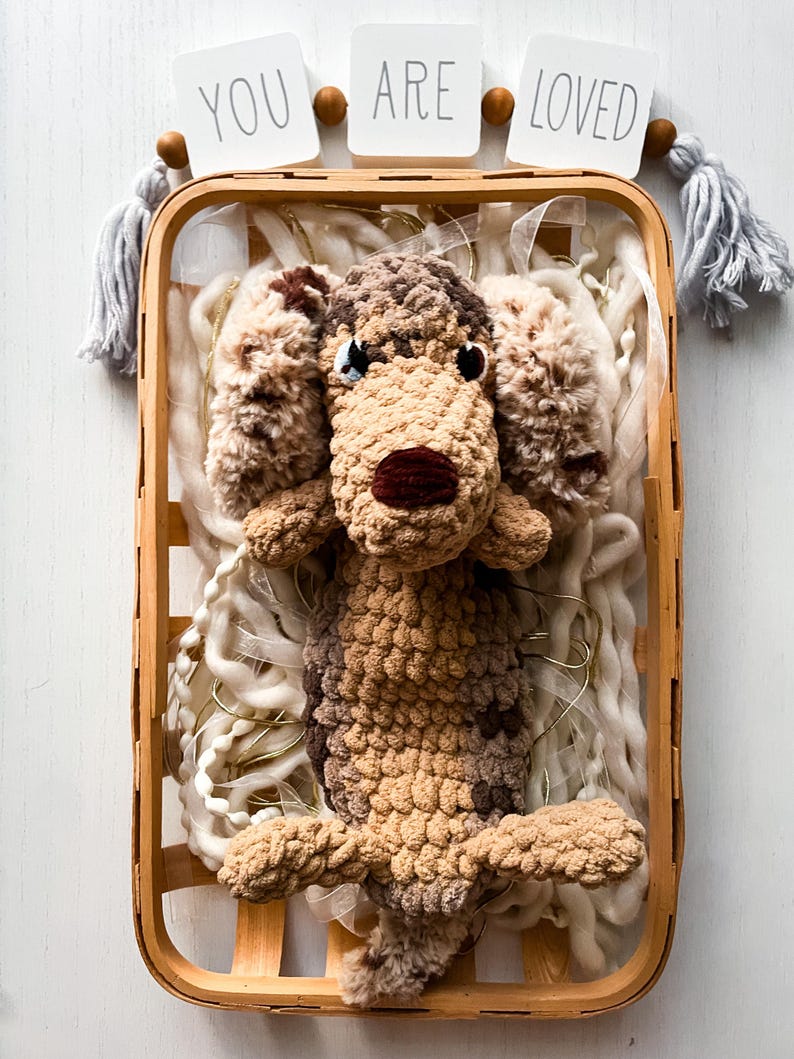May include: A handmade crochet dog toy in a wooden basket, surrounded by white yarn and string lights. The dog is brown and tan with a dark red nose. The words "YOU ARE LOVED" are displayed above the basket.