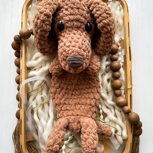 May include: A handmade brown crochet dog toy with black eyes and a brown nose, resting in a wooden basket filled with white yarn and decorative beads. The dog has floppy ears and a textured, plush appearance.