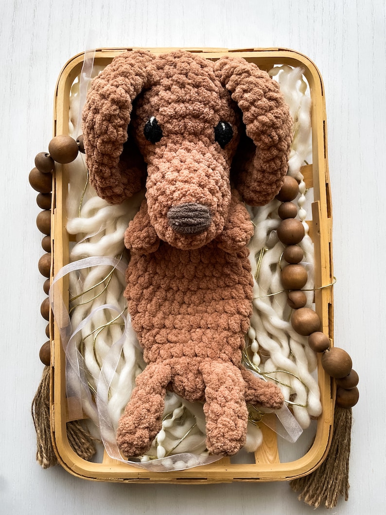 May include: A handmade brown crochet dog toy with black eyes and a brown nose, resting in a wooden basket filled with white yarn and decorative beads. The dog has floppy ears and a textured, plush appearance.