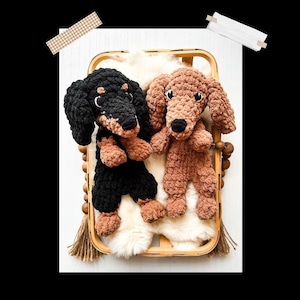 Puede incluir: Dos peluches de dachshund de crochet, uno negro y otro marrón, están sentados en una cesta de madera forrada con piel blanca. La cesta tiene un borla marrón colgando. El texto "DASH THE MINI DACHSHUND" está en la parte inferior de la imagen.