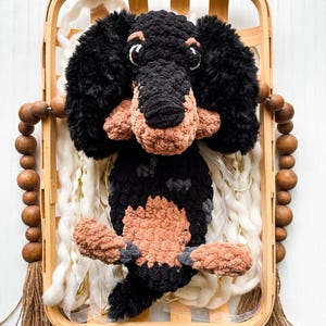 May include: A handmade crochet dachshund dog in a wooden basket. The dog is black and tan with fluffy ears and a long body. The basket has wooden beads and a tassel. The background includes white yarn.