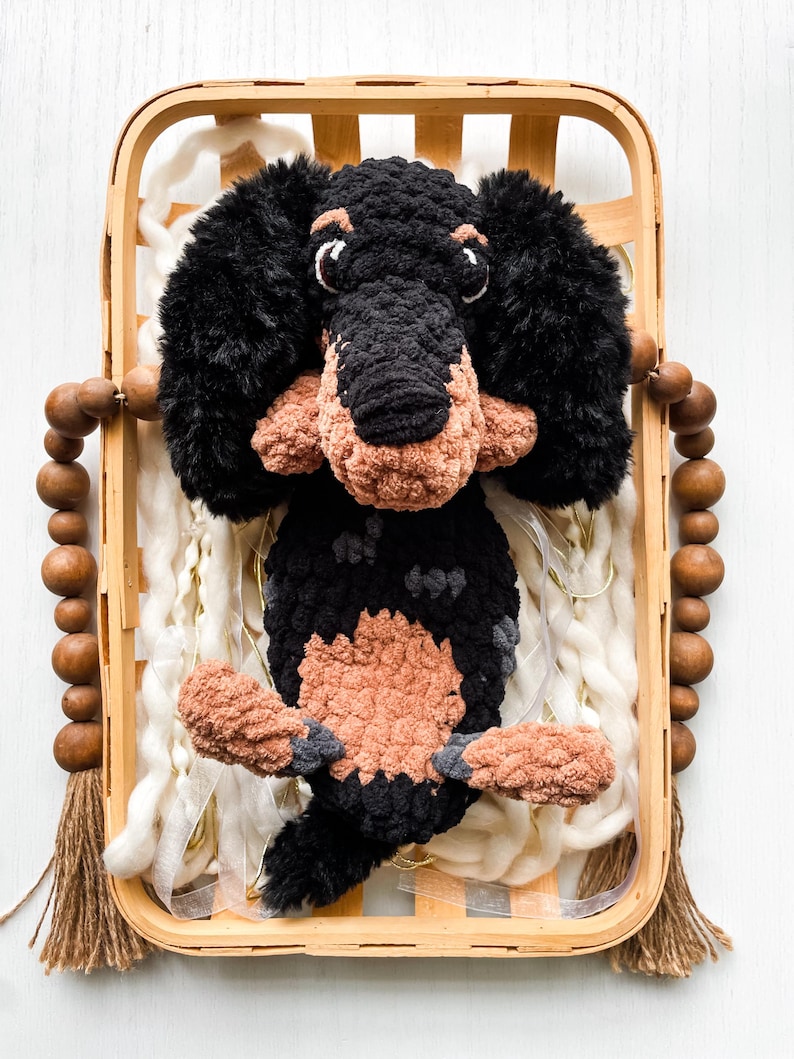 May include: A handmade crochet dachshund dog in a wooden basket. The dog is black and tan with fluffy ears and a long body. The basket has wooden beads and a tassel. The background includes white yarn.