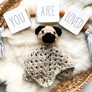 May include: A handmade crochet pug dog security blanket in beige and black, with the words "YOU ARE LOVED" on a banner above. The blanket is on a white faux fur surface, inside a woven basket.