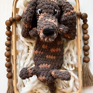 May include: A handmade crochet dog in a wooden basket. The dog is brown with tan markings and has a black nose and white eyes. The basket contains white yarn and is decorated with wooden beads and tassels.