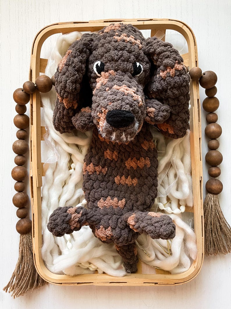 May include: A handmade crochet dog in a wooden basket. The dog is brown with tan markings and has a black nose and white eyes. The basket contains white yarn and is decorated with wooden beads and tassels.