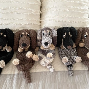 May include: Five handmade crochet dachshund dog stuffed animals in various colors: black, brown, gray, and a mix. Each plush dog has a long body, floppy ears, and small paws. They are arranged on a bed with white pillows.