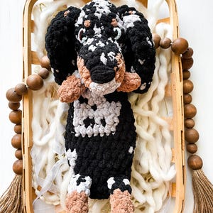 May include: A handmade crochet dog toy with black, white, and tan accents. The dog is in a wooden basket with white fluff and a wooden bead garland. The dog's body is black and white, with tan paws and ears.