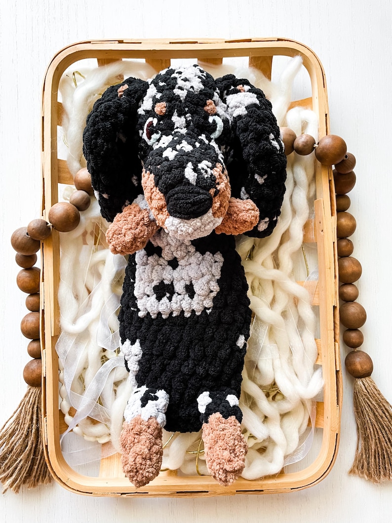 May include: A handmade crochet dog toy with black, white, and tan accents. The dog is in a wooden basket with white fluff and a wooden bead garland. The dog's body is black and white, with tan paws and ears.