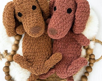 May include: Two handmade crochet plush dogs, one in a medium brown yarn and the other in a dusty rose yarn. Both stuffed animals feature long floppy ears, dark circular eyes, and a small stitched nose, resting on a woven circular tray.