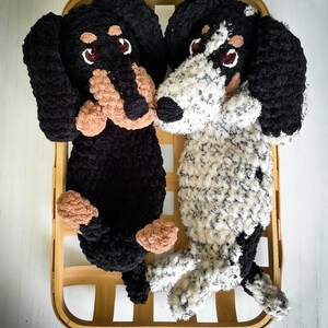 May include: Two plush dachshund dog toys, one black and one white with black spots, rest in a wooden basket. The dogs have brown and tan accents on their faces and paws, with dark eyes.