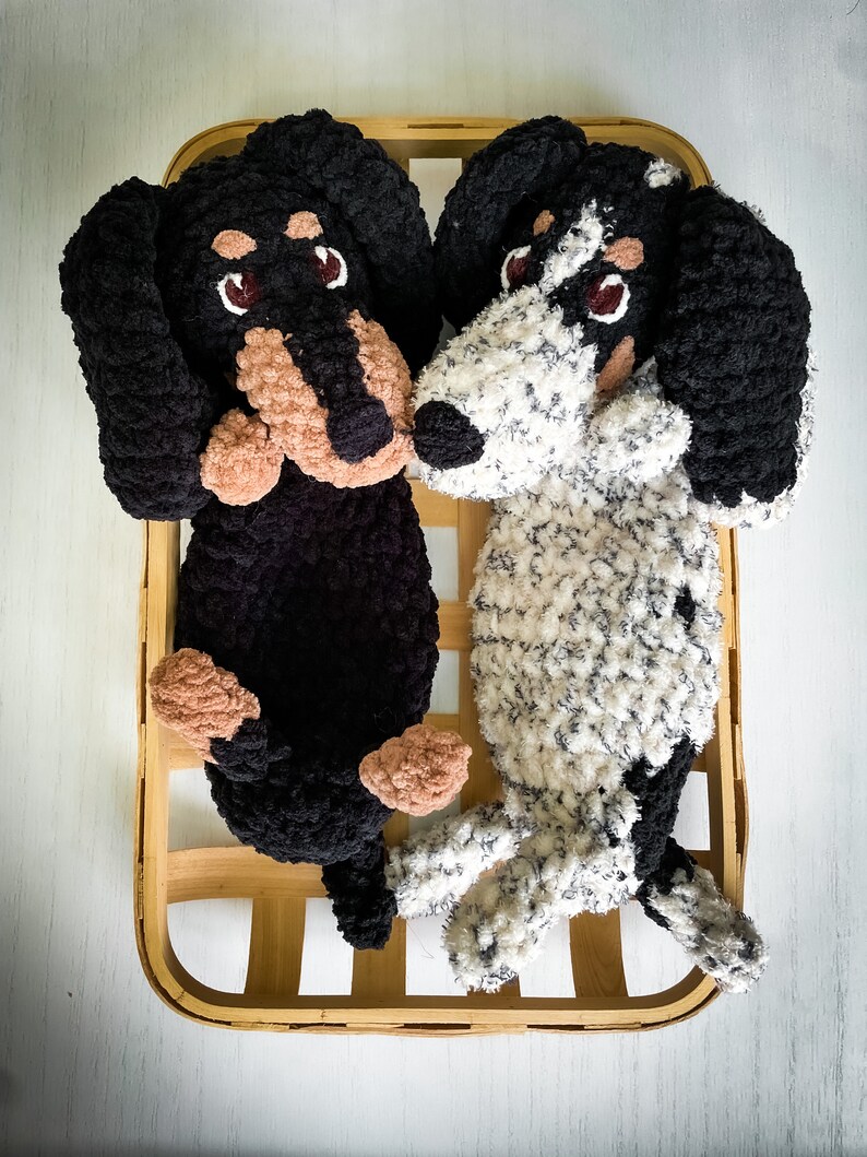 May include: Two plush dachshund dog toys, one black and one white with black spots, rest in a wooden basket. The dogs have brown and tan accents on their faces and paws, with dark eyes.