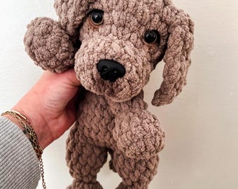 May include: A brown crochet dog plush toy with a textured, chunky yarn finish. The stuffed animal features large, dark, glossy safety eyes, a black plastic nose, and floppy ears. The design is crafted with a soft, looped yarn texture throughout.