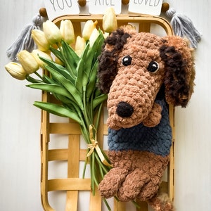 May include: A handmade crochet dachshund dog in a basket with a bouquet of yellow tulips. The dog is brown and wearing a blue sweater. The basket is wooden, and the tulips are tied with a yellow ribbon. The words "YOU ARE LOVED" are displayed above.