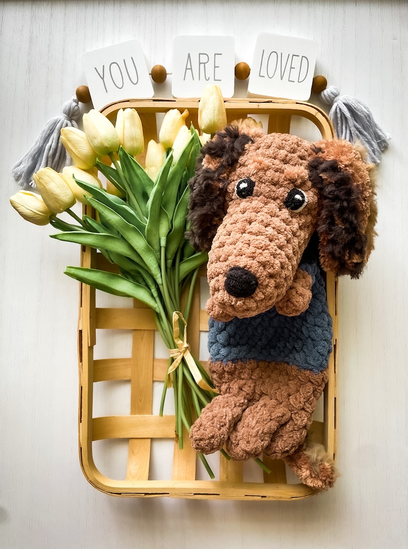 May include: A handmade crochet dachshund dog in a basket with a bouquet of yellow tulips. The dog is brown and wearing a blue sweater. The basket is wooden, and the tulips are tied with a yellow ribbon. The words "YOU ARE LOVED" are displayed above.