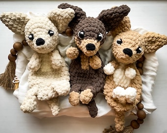 May include: Three crocheted dog plushies in a white basket. The plushies are brown, beige, and white. The plushies have black eyes and noses.