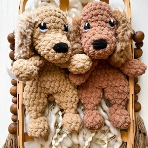 May include: Two handmade crochet dog plushies in a wooden basket. One dog is light brown, the other is a darker brown. Both have black noses and white-rimmed eyes. The basket contains white yarn and is decorated with wooden beads and a tassel.