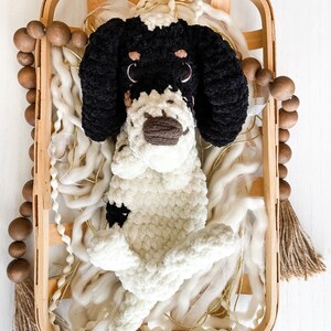 May include: A handmade, plush dog toy in a wooden basket. The dog is black and white, with large floppy ears and a brown nose. The basket is filled with white yarn and gold ribbon. A wooden bead garland and tassel are on the side.