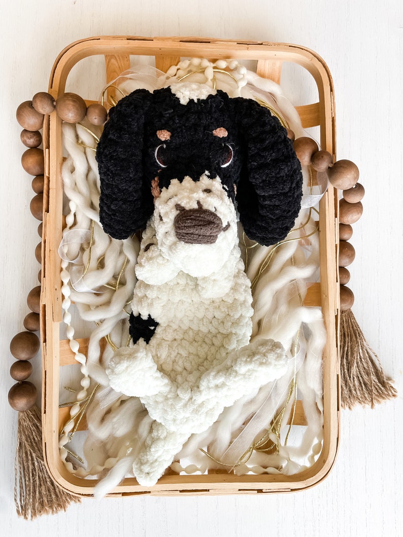 May include: A handmade, plush dog toy in a wooden basket. The dog is black and white, with large floppy ears and a brown nose. The basket is filled with white yarn and gold ribbon. A wooden bead garland and tassel are on the side.