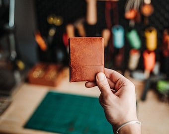 May include: A small, rectangular, brown leather wallet with a smooth, folded flap design. The wallet features a rich, textured surface with visible grain patterns and clean, straight edges, held up against a blurred workshop background.