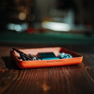 Puede incluir: Una bandeja rectangular de cuero naranja con el texto en relieve "EAGLE SLOW". La bandeja contiene gafas, una pequeña cartera azul y un reloj. La bandeja está sobre una superficie de madera oscura.